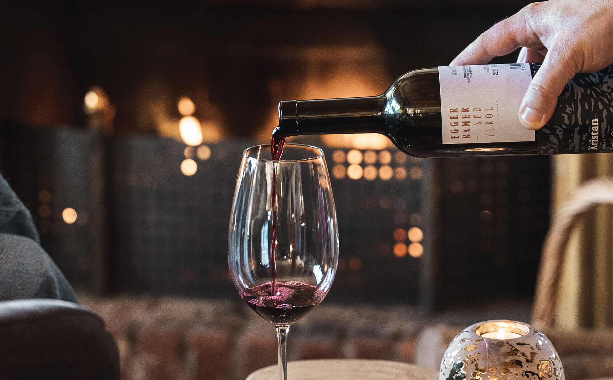 Un bicchiere di vino rosso viene versato, il fuoco aperto brucia dietro di esso - Vitalhotel Erica