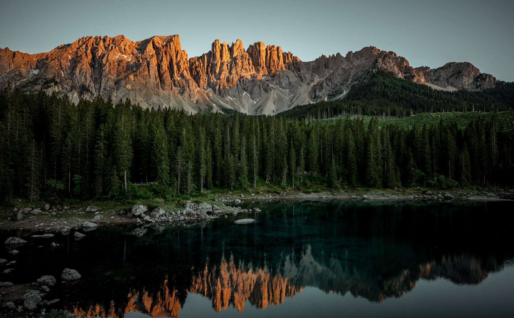 Il lago di Carezza, con il bosco alle spalle e l'area del Catinaccio e Latemar che si illumina di rosso al sole - Vitalhotel Erica