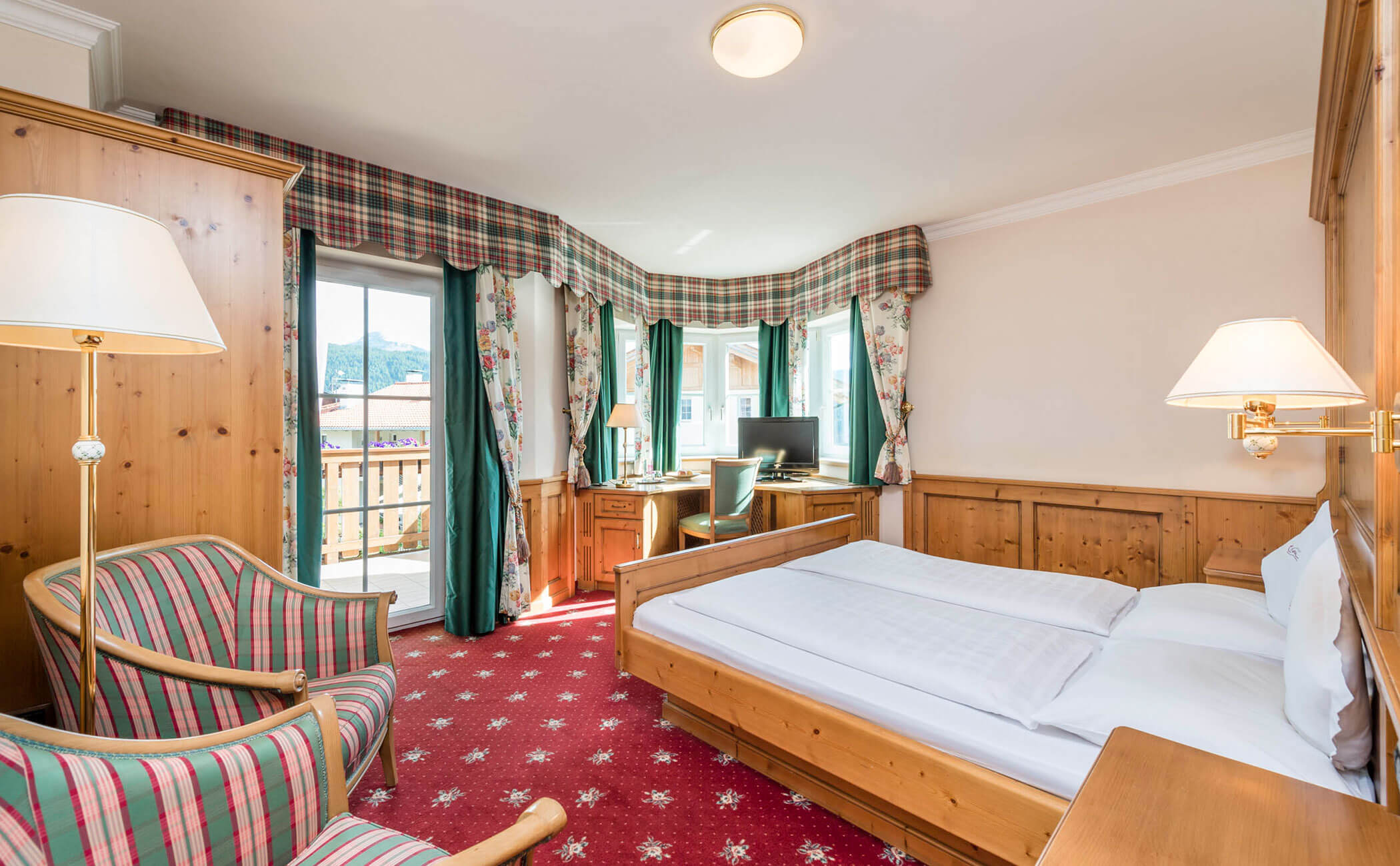 Letto matrimoniale con poltrona e tende rosso-verdi nella camera doppia Latemar - Vitalhotel Erica