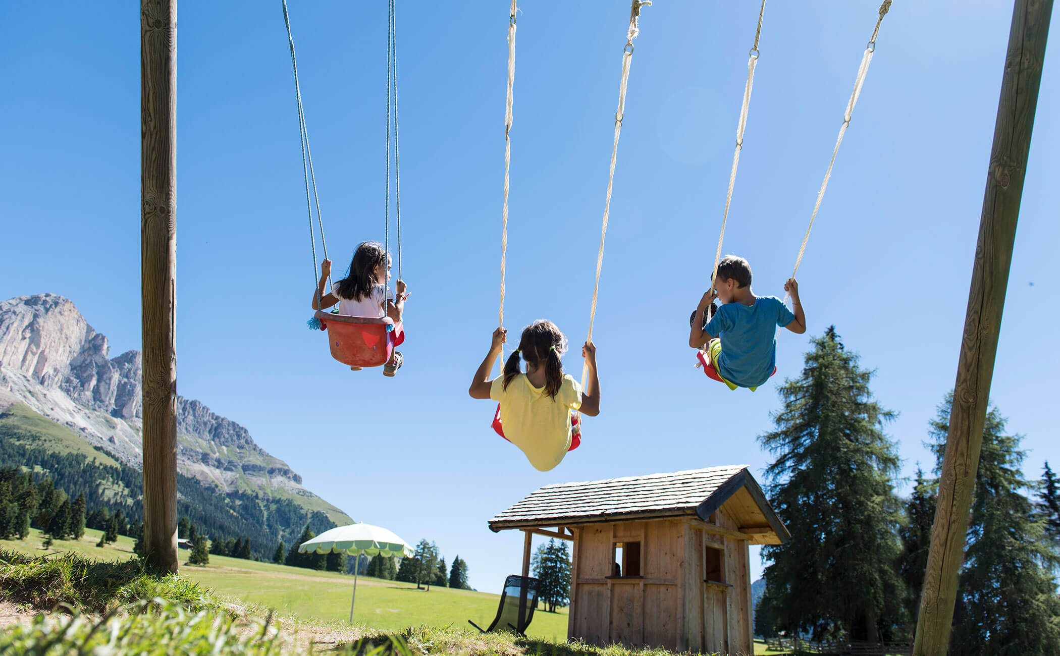 Tre bambini che si dondolano, alle loro spalle una capanna di legno e le cime delle Dolomiti - Vitalhotel Erica