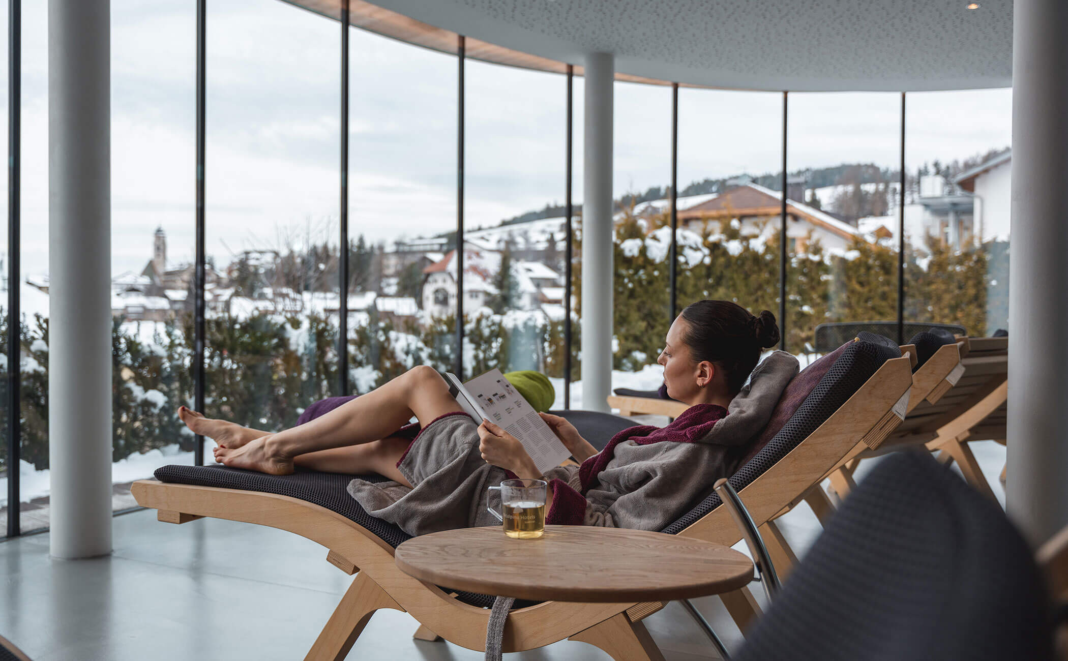 Una donna si sdraia nella sala relax leggendo una rivista, con una tazza di tè accanto - Vitalhotel Erica