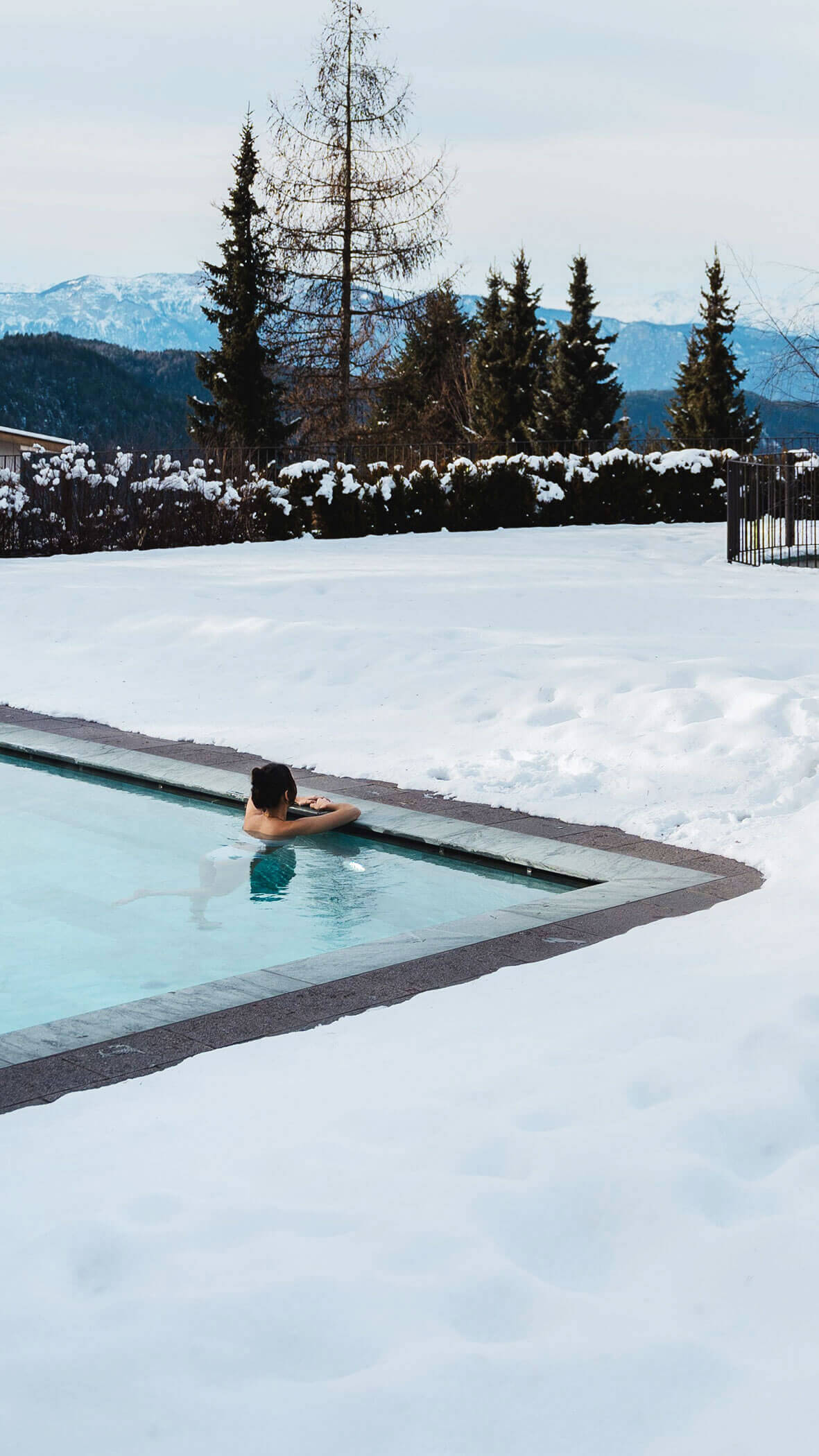 Una donna nella piscina all'aperto guarda verso il villaggio innevato - Vitalhotel Erica