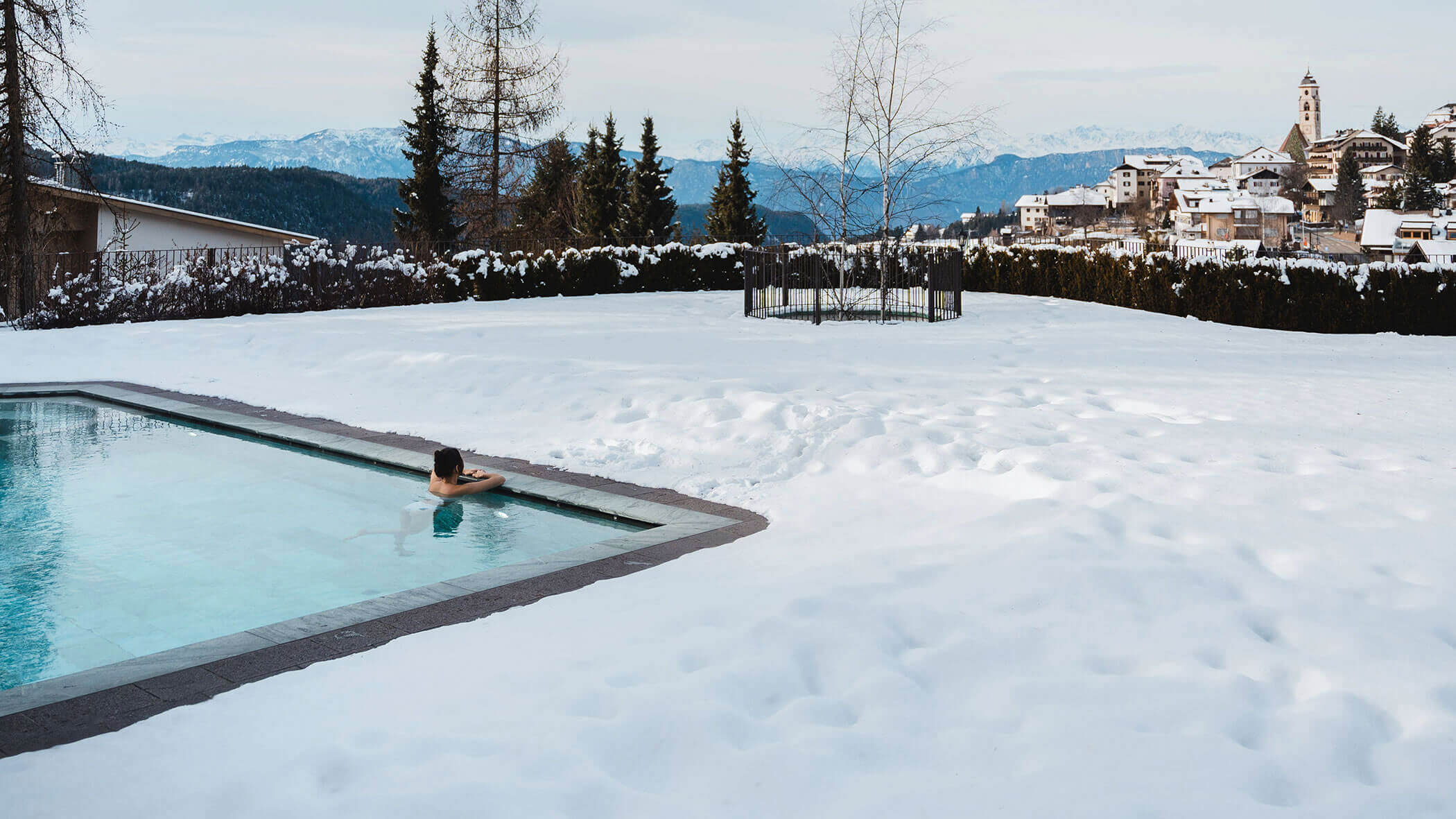 Una donna nella piscina all'aperto guarda verso il villaggio innevato - Vitalhotel Erica