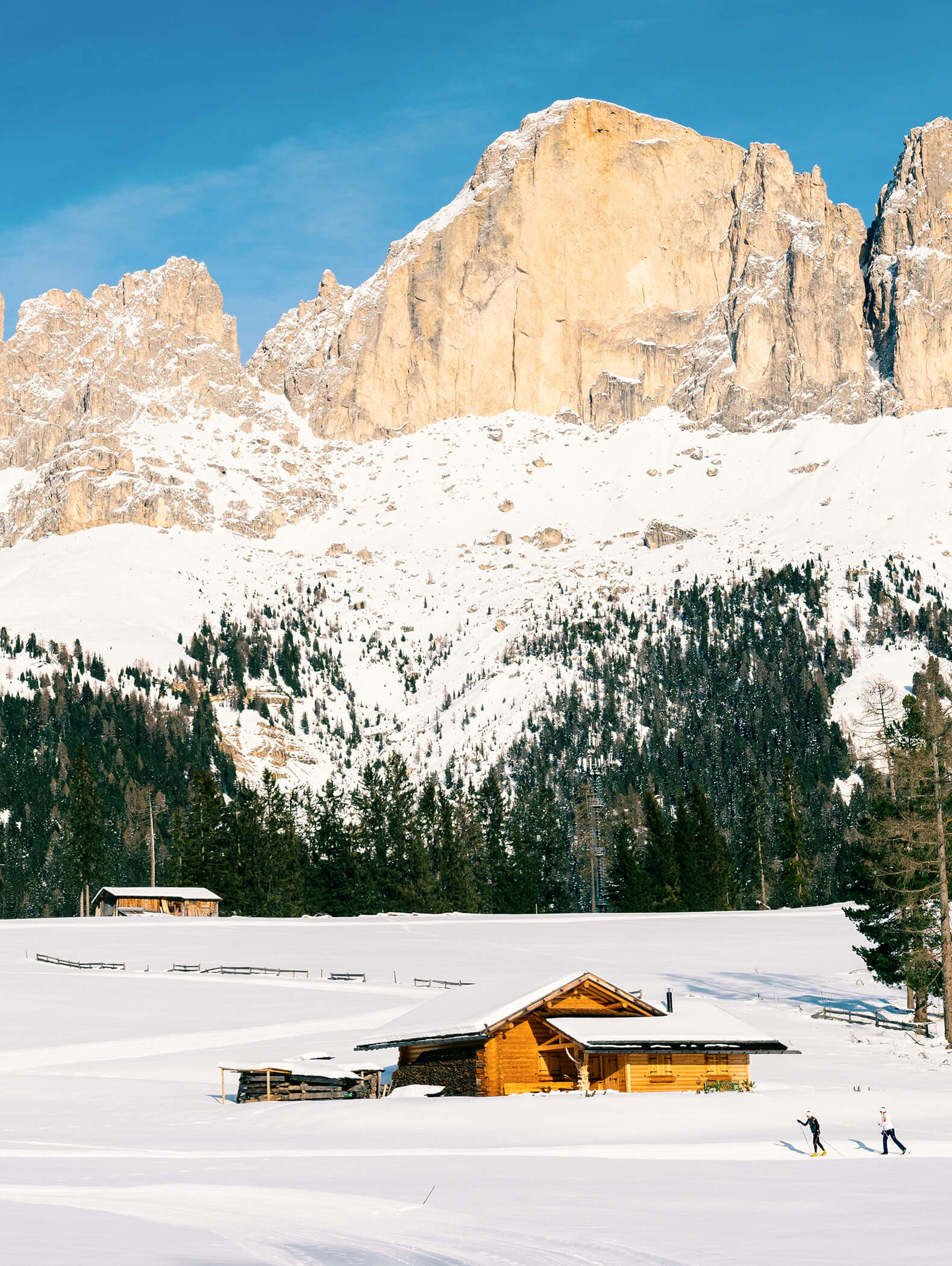 Una baita sopra la pista di fondo in mezzo alla neve, alle spalle alcune cime di montagna - Vitalhotel Erica