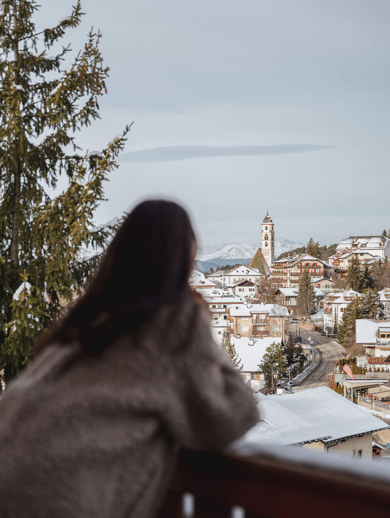 Eine Frau steht auf dem Balkon und blickt runter ins verschneite Dorf - Vitalhotel Erica