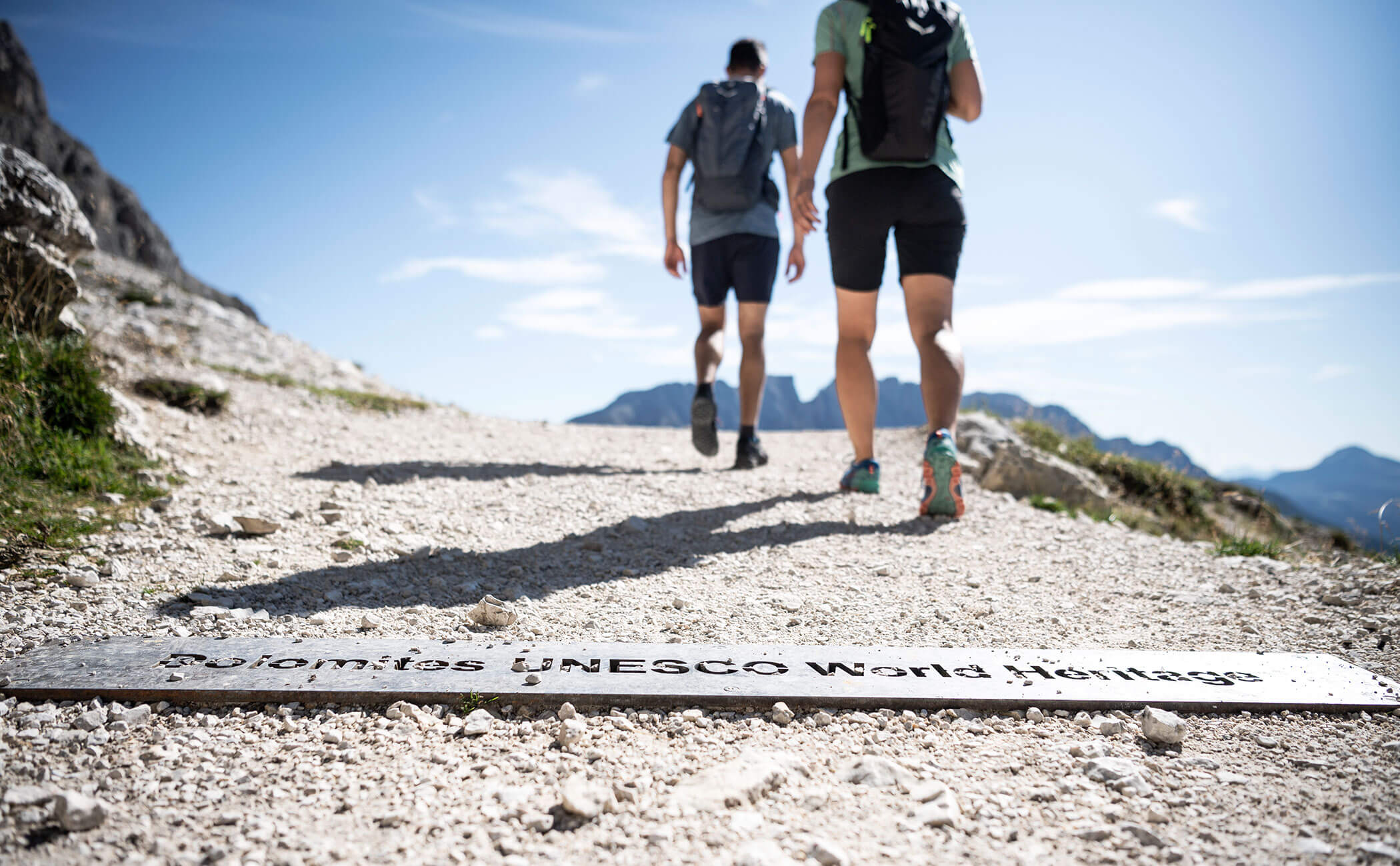 Due escursionisti in cammino. Un cartello sul sentiero ghiaioso con la scritta Dolomiti Patrimonio Mondiale UNESCO - Vitalhotel Erica