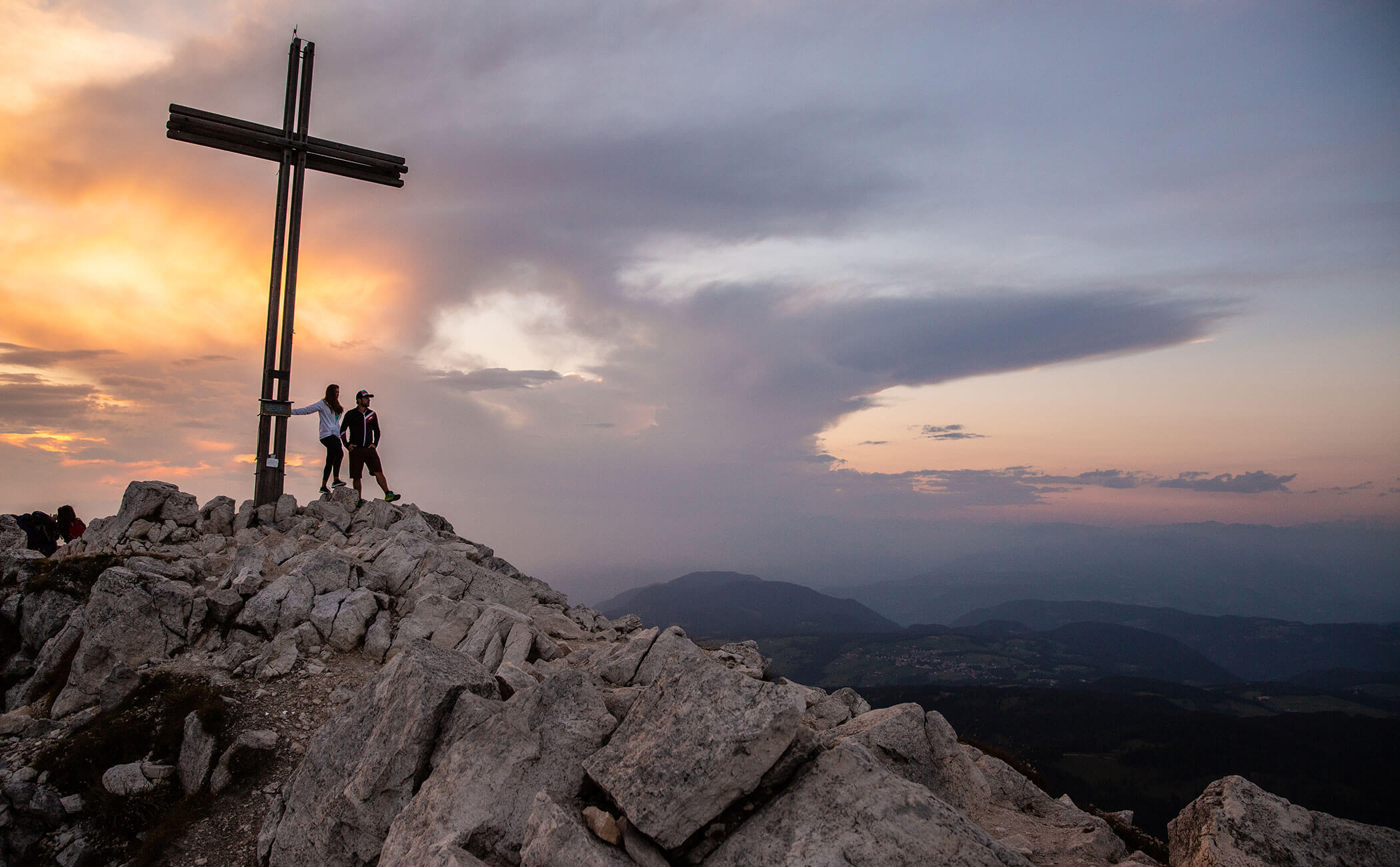 Una coppia in piedi sulla croce di vetta del Weisshorn e guarda in lontananza, con il sole che tramonta alle loro spalle - Vitalhotel Erica