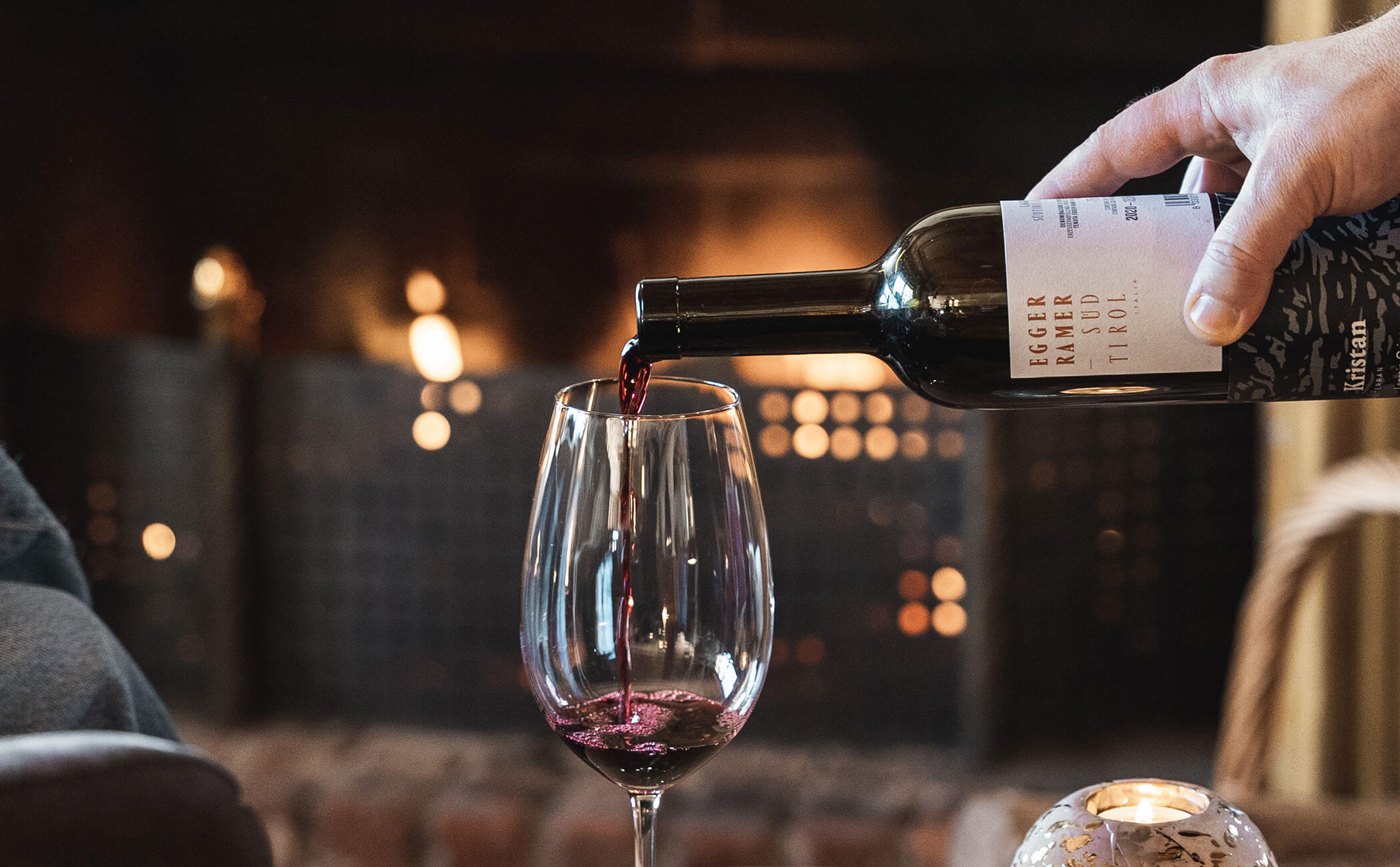 Un bicchiere di vino rosso viene versato, il fuoco aperto brucia dietro di esso - Vitalhotel Erica