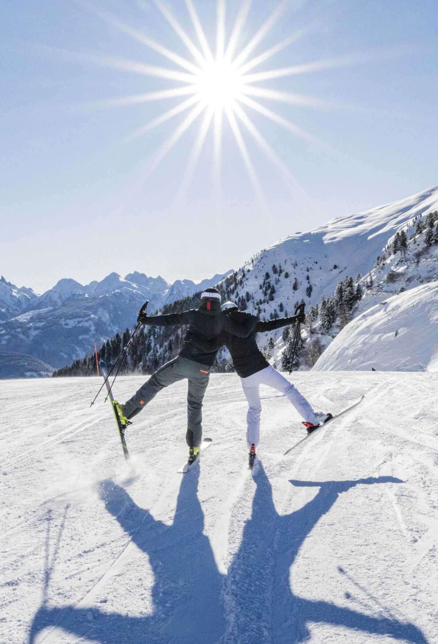 Zwei Skifahrer, die sich umarmen, heben auf einem verschneiten Hang jeweils ein Bein in die Höhe. - Vitalhotel Erica